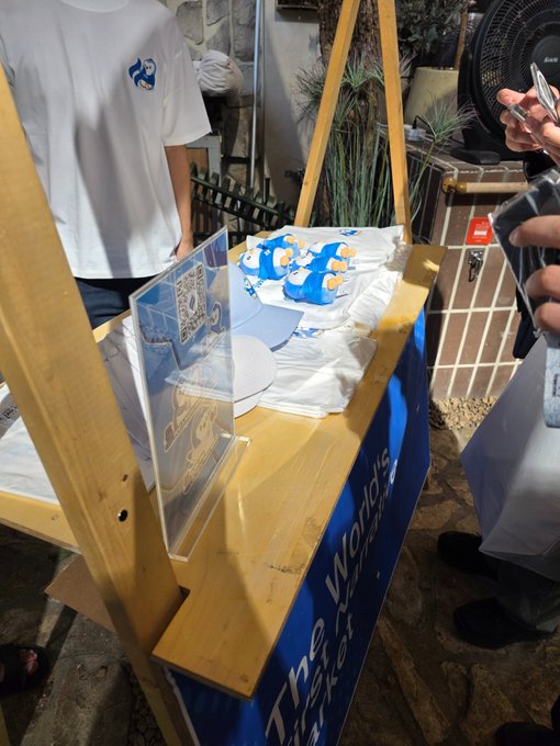 The image shows a booth featuring Pudgy Penguins merchandise, with a display table bearing the text "...gle's World" (likely "Pudgy's World") and a cute penguin mascot holding a QR code sign, while attendees in white t-shirts stand nearby at the event. This matches the post's caption about attending the Huddle at KBW with the Pudgy Penguins community, showing the penguin-themed booth setup that brought the narrative-themed event together.