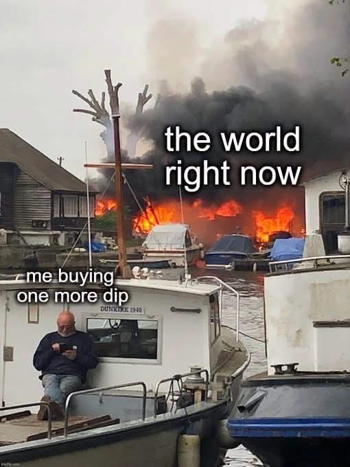 A meme photograph showing a man sitting on a boat calmly looking at his phone while a large fire blazes in the background; text reads 'the world right now' pointing to the fire and 'me buying one more dip' pointing to the man.