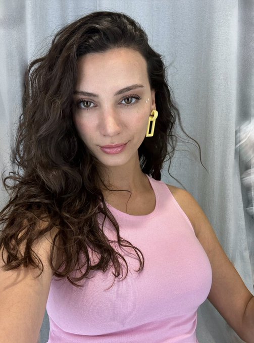 Selfie of a young woman with long curly brown hair wearing a pink tank top and yellow earrings against a light curtain background.