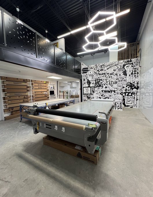 An industrial workshop space featuring a long roll-to-roll printer machine in the center, geometric hexagonal LED ceiling lights, and a black-and-white mural wall with text including 'Sevilla' and 'Cristina'.