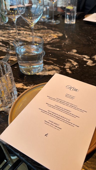 A restaurant table setting with a printed menu from "Il Cun" featuring items like Sea Jackfruit and Niçoise Gravlax, accompanied by wine glasses and water tumblers on a dark marble surface.