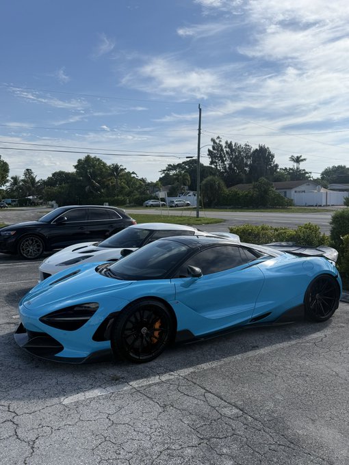 The image shows a blue sports car parked in a parking lot, with another car behind it. There are also two people visible near the cars. The caption suggests that someone might be considering selling the blue sports car for Bitcoin, but there isn't enough information provided about whether this is actually happening or not.