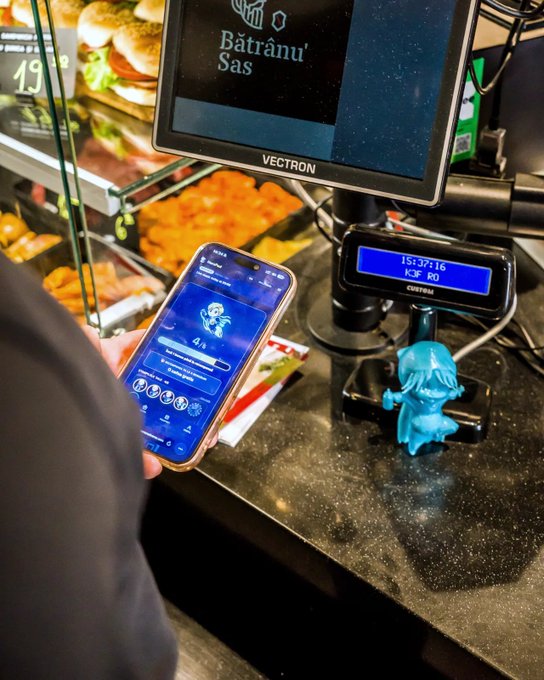 A person uses a smartphone displaying a loyalty app at a checkout counter featuring a Vectron POS system labeled 'Bătrânu Sasu' and 'K3F R0' (15:37:16), with a small blue figurine resting on the counter.