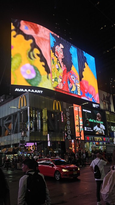 A nighttime street scene in Times Square features large illuminated billboards, including one displaying artwork with the text 'AVAAAAAART' and McDonald's and Forever 21 storefronts below.