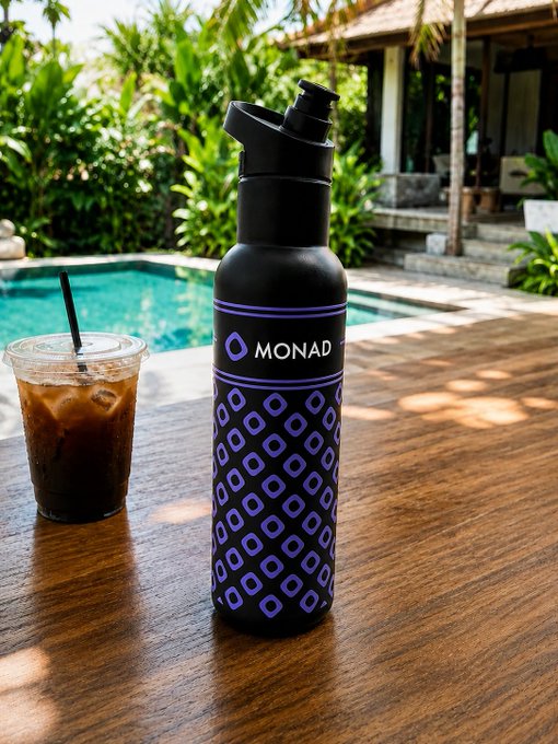 A black MONAD-branded water bottle with purple geometric patterns and a spout lid sits on a wooden table beside an iced coffee, with a swimming pool and tropical setting in the background.