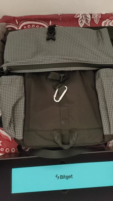 A gray and olive green checkered backpack with black accents and a carabiner clip sits on a bed, with a teal Bitget box visible at the bottom of the frame.
