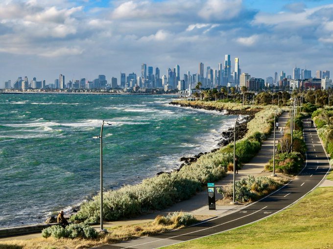 A scenic photograph of a coastal bike path running alongside the ocean with a city skyline visible in the distance under a partly cloudy sky.