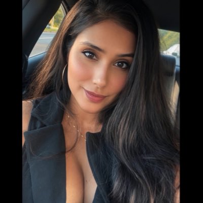 A selfie of a young woman with long dark hair wearing a black top and necklace, taken inside a vehicle.
