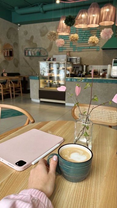 A hand reaches for a blue ceramic mug of latte art coffee on a wooden café table, with a pink laptop, glass vase of flowers, and pendant lamps visible in the background.