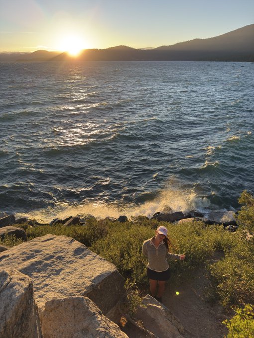 A woman in a pink cap and dark shorts hikes along a rocky lakeshore trail at sunset, with mountains visible across the water.