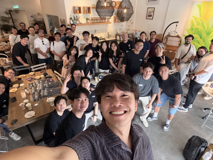 A large, smiling group poses for a selfie-style group photo inside a bright café, gathered around wooden tables and looking up at the camera with cheerful expressions. The casual, intimate setting and post text suggest this is a cozy founders' hangout or appreciation event for early supporters, power users, and partners, with cofounder @cho_dominic appearing to have shared updates during the gathering.