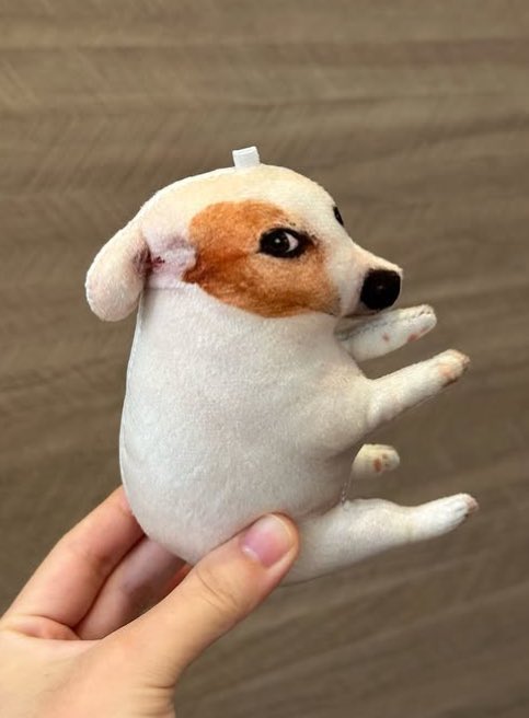 A small plush dog with tan and white fur is held up by a hand against a wooden background, appearing to be a prototype or sample of an upcoming $SDOG plushie release.