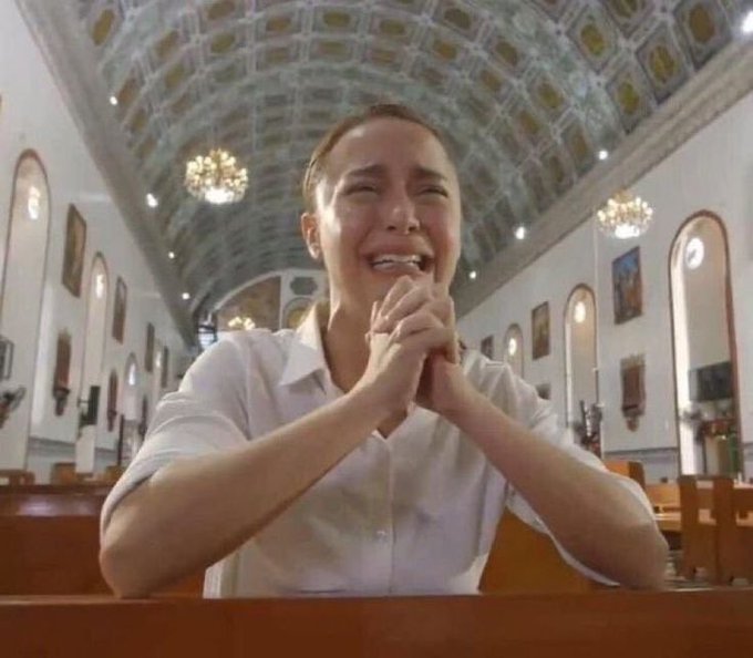 Photo of a young woman in a white shirt sitting in a church pew, hands clasped together, crying and smiling with visible tears, under an ornate painted ceiling with chandeliers.