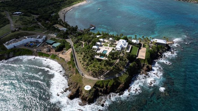 An aerial view shows a luxurious coastal estate situated on a rocky peninsula surrounded by clear turquoise waters and crashing waves.