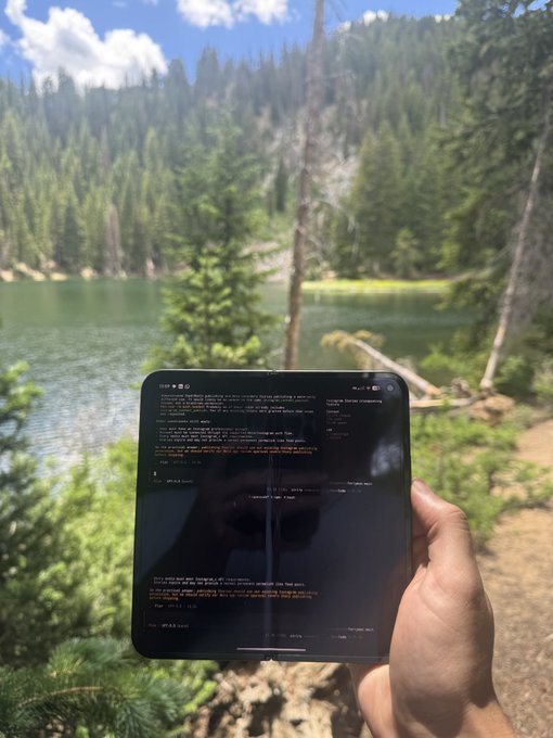 A hand holds a tablet displaying code with orange syntax highlighting against a dark theme, set against a serene backdrop of a forest lake with evergreen trees reflecting in the calm water. The scene visually reinforces the post's theme of remote, nature-immersed software development and income generation with ferryman.io and opencode on a phone.