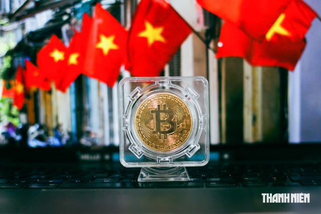 A physical Bitcoin coin sits in a clear acrylic display case on a laptop keyboard, with multiple Vietnamese flags (red with yellow star) in the background. The watermark 'THANH NIÊN' appears in the bottom right corner.