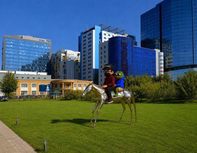 Two children wearing masks ride a shiny silver metallic horse sculpture on a grassy lawn, with modern glass and blue high-rise buildings in the background.