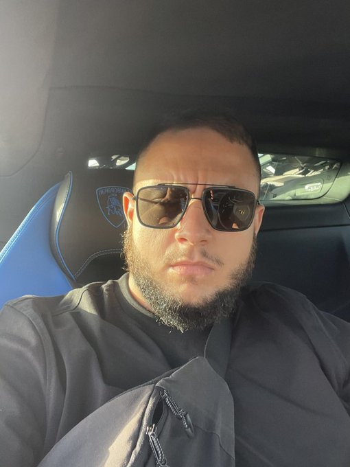 A selfie of a bearded man wearing dark sunglasses and a black shirt, sitting in the driver's seat of a vehicle with a blue racing-style seat featuring a lion logo in the background.