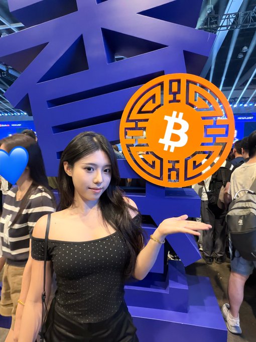 A young woman in a black polka-dot top poses at what appears to be a crypto conference, holding up her hand toward a large orange Bitcoin logo against a blue backdrop.