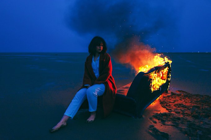 A woman in a red coat and white pants sits on a burning chair on a beach at dusk, with smoke rising into the deep blue sky.