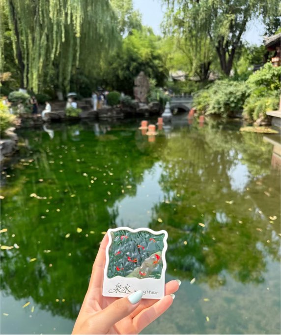 A hand holds a sticker labeled '泉 Water' depicting fish swimming among aquatic plants, held in front of a serene Chinese garden pond with weeping willows and a stone bridge.