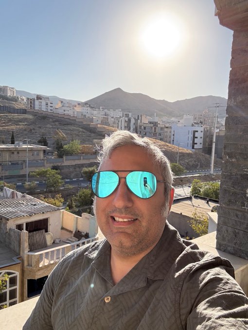 A selfie of a smiling man with short gray hair and reflective blue sunglasses, with a sunny landscape of buildings and mountains in the background.