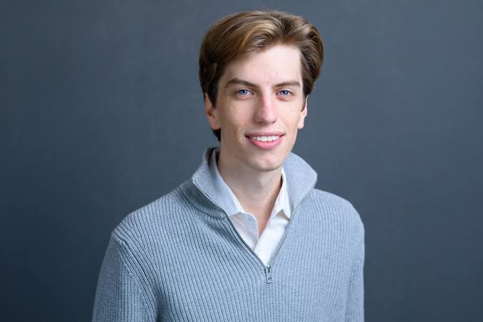 A young man with brown hair and blue eyes smiles at the camera while wearing a gray quarter-zip sweater over a white collared shirt, posed against a plain gray background. The image appears to be a professional portrait of Leopold Aschenbrenner, referenced in the post regarding a rug-pull scheme affecting his retail trading audience.