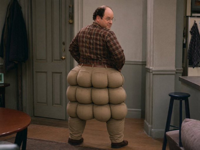 A man with an exaggeratedly large, padded backside stands looking over his shoulder in a doorway, wearing a plaid shirt and glasses. The image humorously illustrates the post's joke about stubbornly insisting "BITCOIN BOTTOM IS IN" over and over, as the character's rear is comically inflated like an "ASS-ET" refusing to deflate.