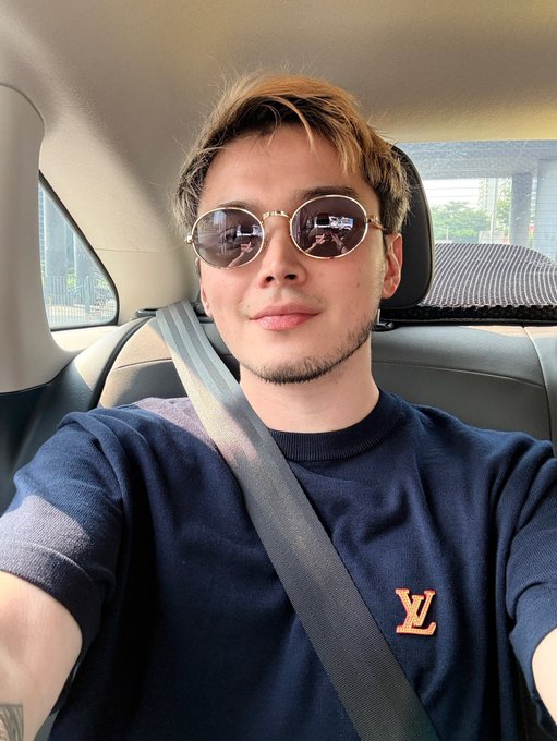 A young man with blonde-highlighted hair wears round sunglasses and a navy blue sweater with a Louis Vuitton "LV" logo while sitting in a car wearing a seatbelt.