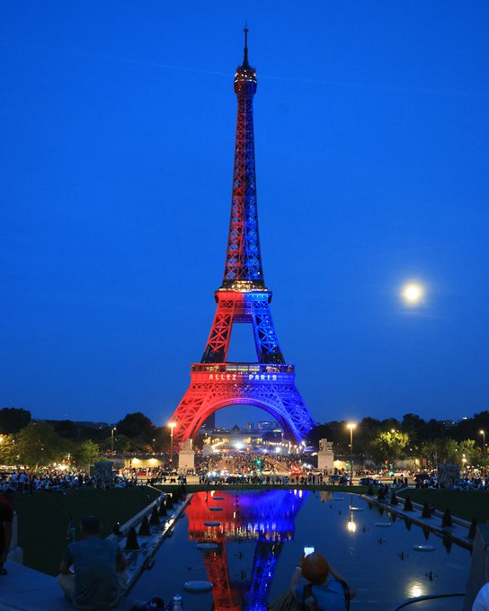 A nighttime photo of the Eiffel Tower, taken by people sitting near a pool that reflects the tower's lights from below. The night sky provides an enchanting background for the scene.