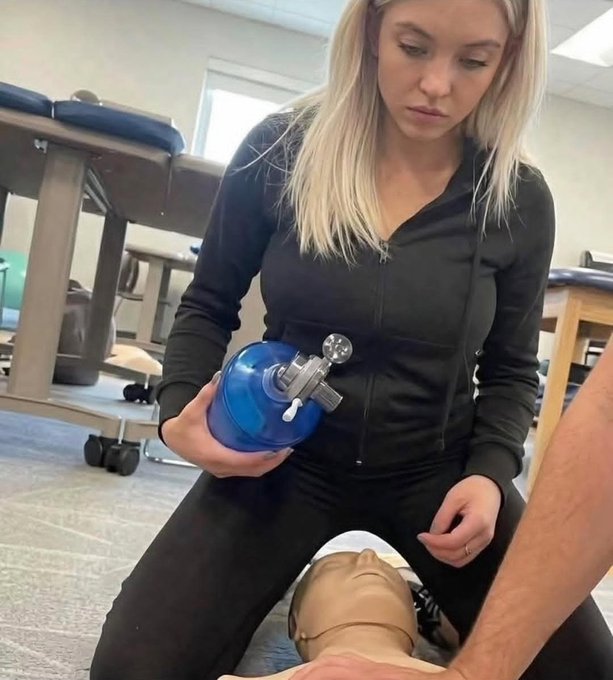A blonde woman in a black tracksuit practices administering CPR using a bag-valve mask resuscitator on a training mannequin in a clinical classroom. The image humorously depicts the post's fictional scenario of Sydney Sweeney attempting CPR on the poster, showing the proper technique she likely failed to use.