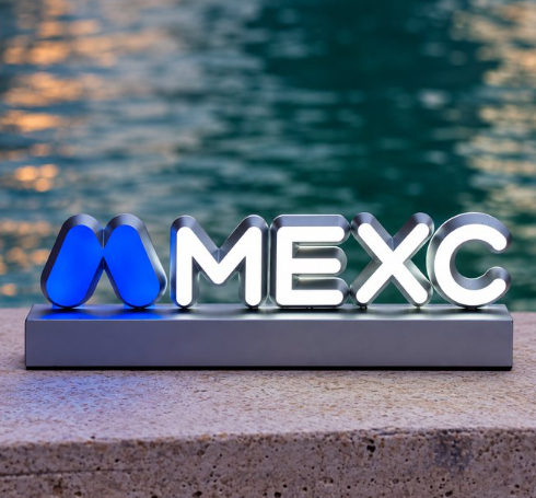 The image shows an illuminated "MEXC" sign/logo placed on a ledge by water at dusk, with the platform's name prominently displayed in white lettering alongside its blue M-shaped logo. It visually anchors the post's discussion about MEXC's unique approach to offering stock exposure on a centralized exchange, emphasizing the brand at the center of this financial conversation.