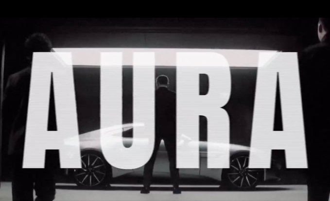 A cinematic image showing three men in suits standing in a garage with a sleek car, with the large white text 'AURA' overlaid across the scene.
