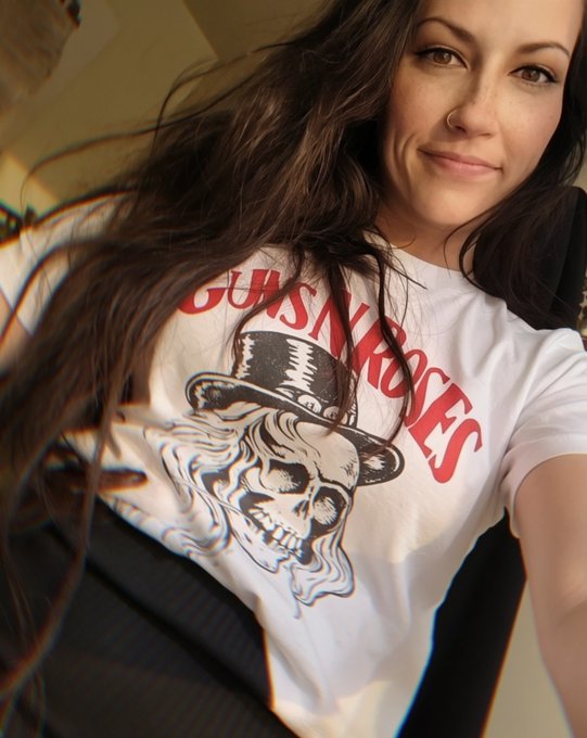 A selfie of a woman with long dark hair wearing a white Guns N' Roses t-shirt featuring the iconic skull with top hat logo.