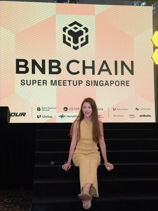 A woman in a yellow dress poses in front of a "BNB Chain Super Meetup Singapore" banner, surrounded by sponsor logos including Aster, Lista DAO, and Unitus. The image reflects the post's description of the event's signature yellow theme and BNB Chain branding in Singapore.