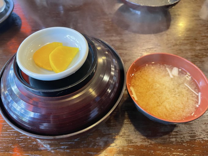 A wooden table displaying a small white dish with yellow pickled vegetables on top of stacked black and purple lacquered bowls, next to a red-rimmed bowl containing a clear broth.