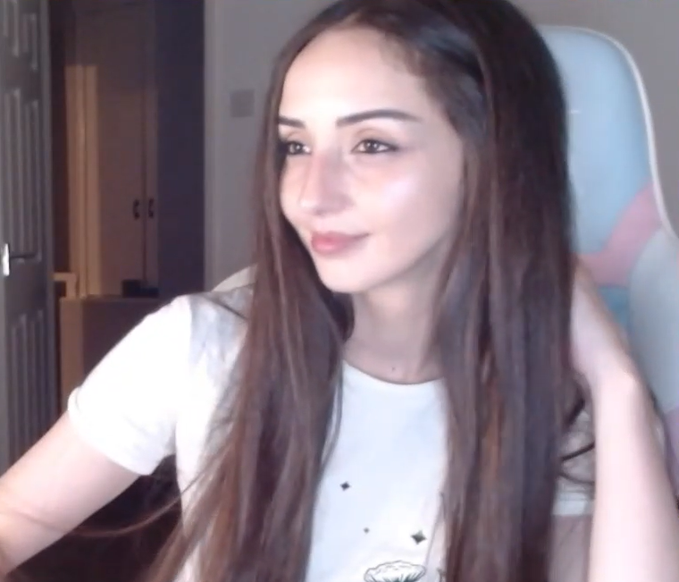 A woman with long brown hair, wearing a white t-shirt, smiles at the camera while sitting in what appears to be a gaming chair.