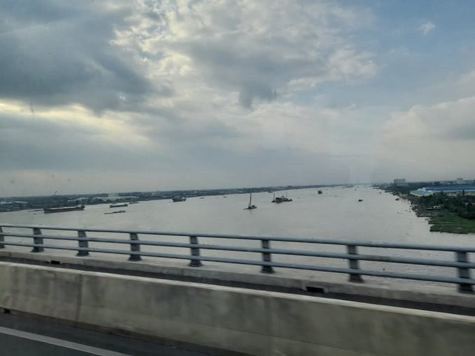 A photograph taken from a bridge overlooking a wide river with several boats and ships, under a cloudy sky, with a concrete highway barrier in the foreground.