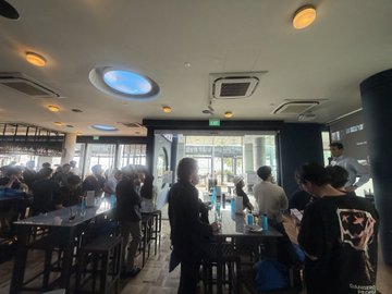 An interior view of a dimly lit restaurant or café with a distinctive circular blue light fixture on the ceiling, showing several patrons seated at high-top tables and at the bar counter.