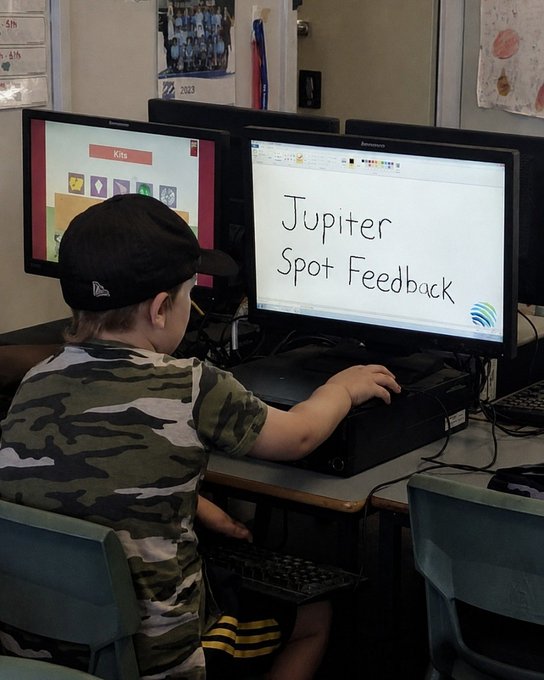 A young student in a camouflage shirt and black cap sitting at a dual-monitor computer station; the main screen displays handwritten text reading 'Jupiter Spot Feedback'.
