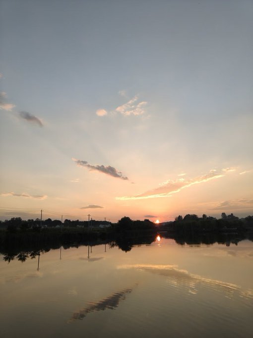 A serene sunset over a calm lake with the sun low on the horizon, casting orange and pink hues across the sky and water, with silhouettes of trees and power lines in the distance.