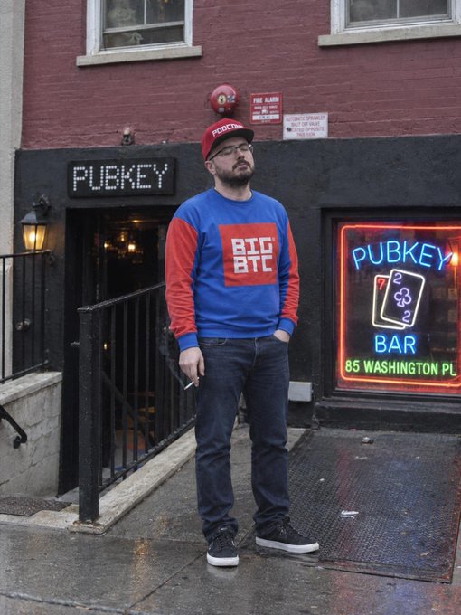 A bearded man wearing a red "Pubkey" cap and a blue sweatshirt with "BIG BITE" printed on it stands on a wet sidewalk outside the Pubkey Bar at 85 Washington Pl.