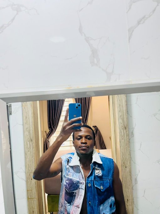 A mirror selfie shows a man holding a blue smartphone, wearing a distressed sleeveless denim jacket, standing in a room with white marble-patterned walls and curtains visible in the reflection.