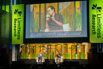 A conference stage for 'Limitless Research' featuring two people seated on stage with a large screen showing a speaker against a yellow floral background.