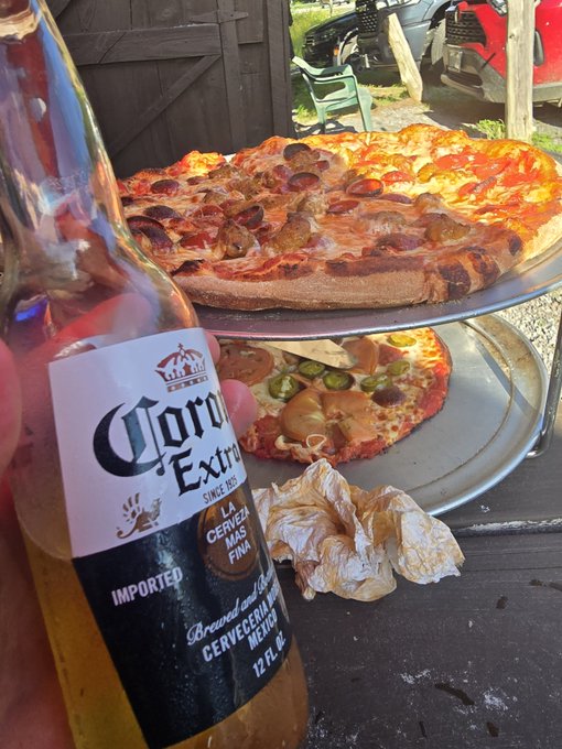 A hand holds a bottle of Corona Extra beer in the foreground, with two pizzas on metal trays (one topped with sausage and pepperoni, the other with tomatoes and jalapeños) resting on a wooden table behind it.