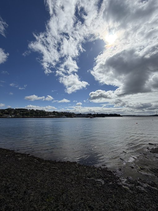 A landscape photograph of a calm body of water under a dramatic sky with scattered clouds, with the sun shining brightly through the clouds and a small shoreline visible in the distance.
