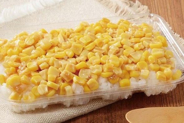 A clear plastic container filled with white rice topped with yellow corn kernels and pieces of egg yolk.