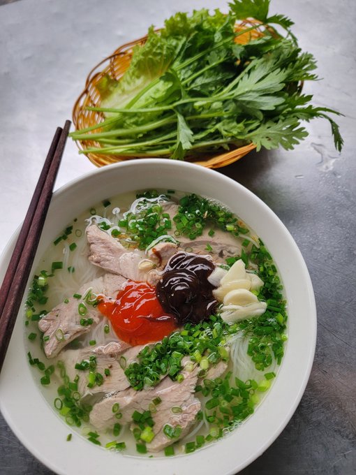 A bowl of Vietnamese-style noodle soup with sliced pork, green onions, chili sauce, and hoisin sauce, served with a side basket of fresh herbs and lettuce.