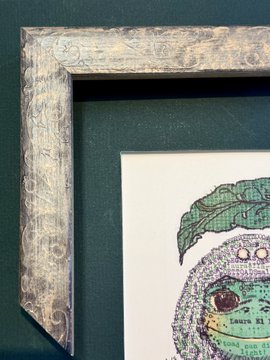 A close-up of the top-left corner of an ornate silver-toned frame with decorative scrollwork, showcasing a partial view of a green-toned artwork featuring a leafy branch and the visible text "Laura E" and "TEEN P."—related to the post about framing artwork by @iamlaurael.