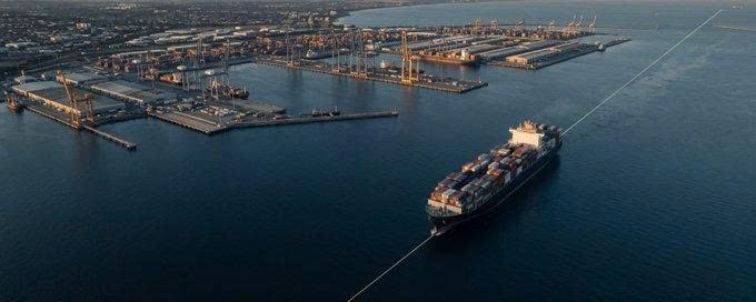 An aerial view of a container ship leaving a large port facility filled with shipping containers and cranes.